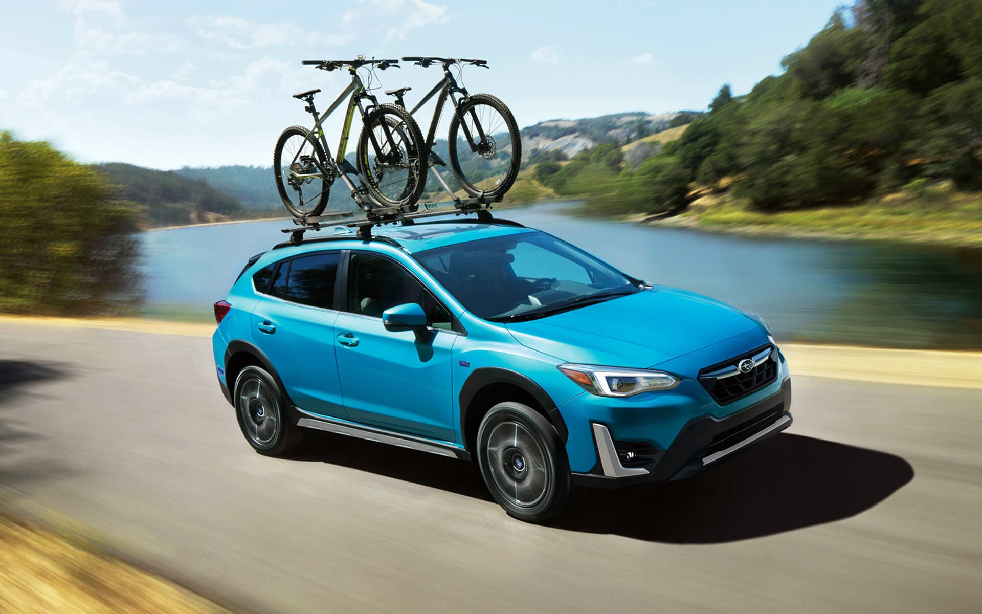 A blue Crosstrek Hybrid with two bicycles on its roof rack driving beside a river | Sunset Hills Subaru in Sunset Hills MO