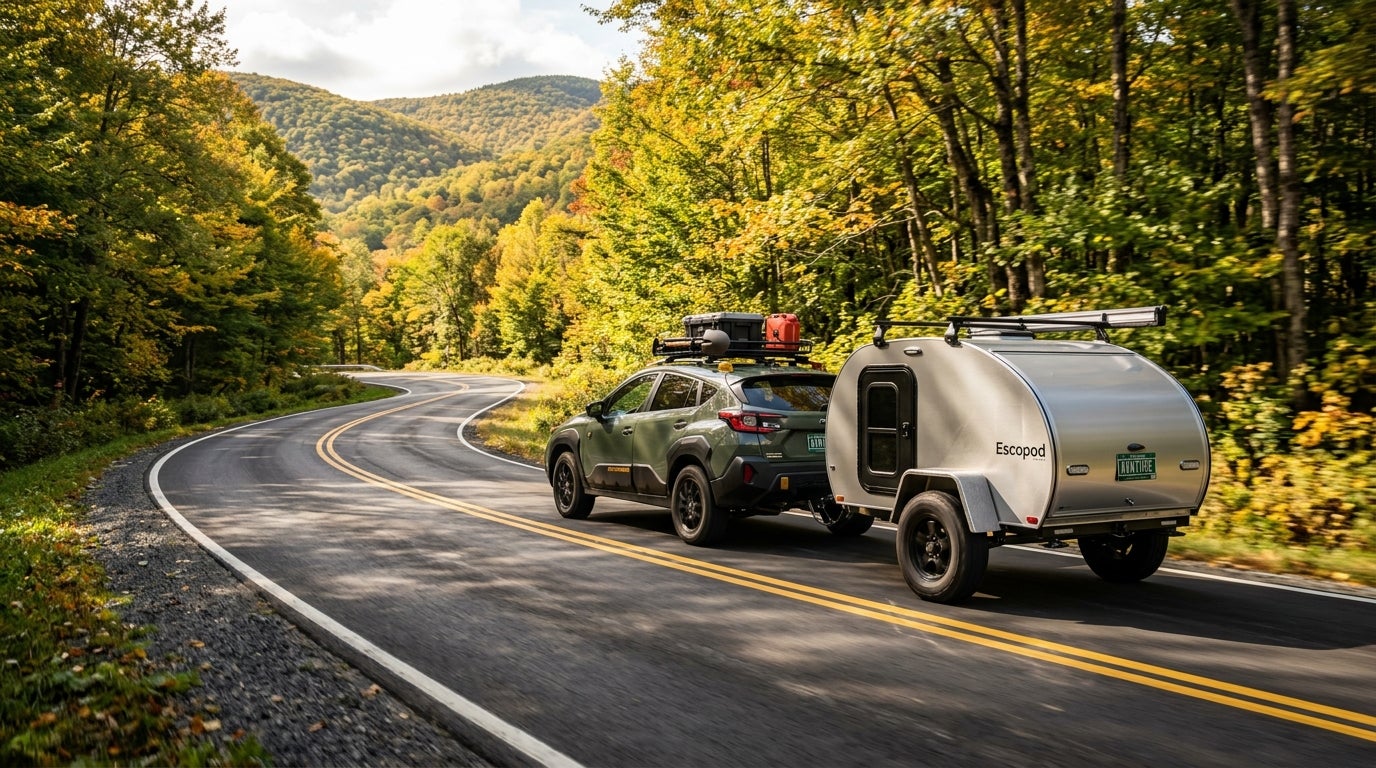 Olive green SUV towing a silver Escopod teardrop camper along a winding mountain road framed by fall foliage. (Representative image for illustration purposes only. Actual vehicle may vary based on trim and configuration.)